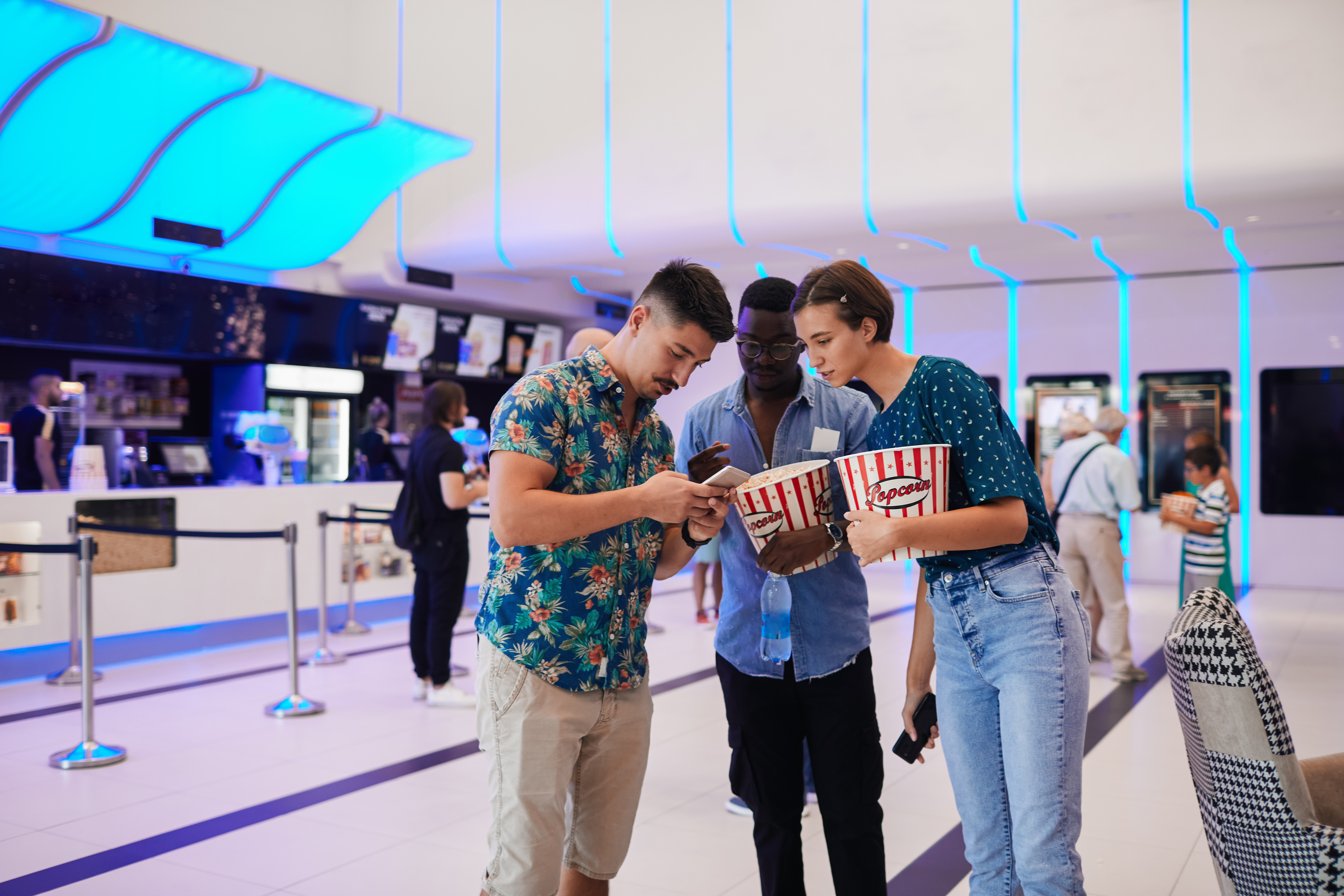 Three people at a movie theater, with two holding popcorn, all leaning over one phone