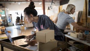 Two women in warehouse, with woman in forefront leaning over her computer