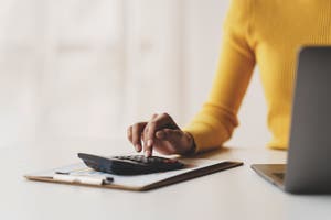 A woman is focused on using a calculator while seated at a desk, surrounded by office supplies and paperwork.