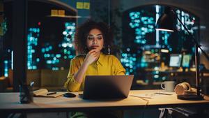 woman sitting at night in an office in front of her computer thinking with tons of other office lights from different buildings illuminating the window. 