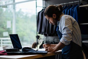 Businessman in a retail suit shop writing in a notebook with a laptop open.