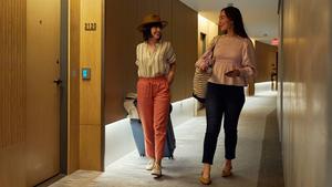 A woman with a hat and pulling luggage walks down the hallway of a hotel with a woman in a pink shirt holding a phone.