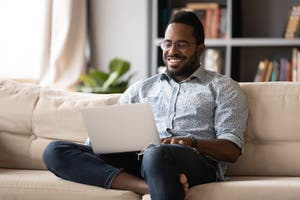 A man sits on his couch and smiles while typing on his laptop.