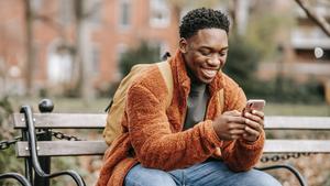 A man sits in a park and smiles at his phone.