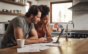 A couple sits at their kitchen counter and reviews emergency savings on their laptop.