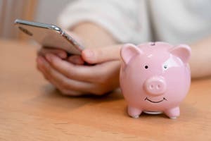 A man types on his phone next to a piggy bank.