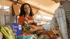 Mujer sonriente en su puesto del mercado de agricultores con un signo de código QR de PayPal, con cliente escaneando el código QR en su teléfono