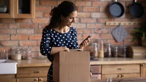 Woman in kitchen holding cell phone and unwrapping a cardboard box package