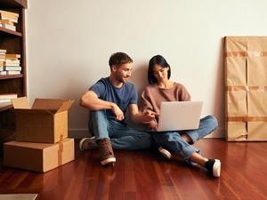 A couple are sitting on the floor of their new apartment and looking at the laptop together