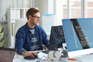Man in glasses and a denim shirt doing IT development work at a desktop computer