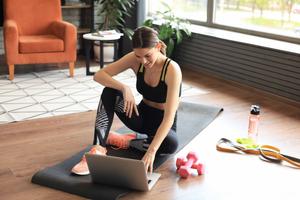 Woman in a black workout set and orange sneakers sitiing on a yoga mat on the floor, using her laptop to adapt to changing clientele needs