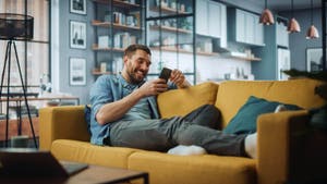 Homme en chemise en jean se prélassant sur un canapé jaune et souriant à son smartphone
