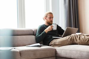 Man sitting on the couch at home with a laptop and a cup surrounded by paperwork