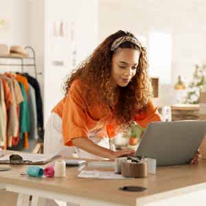 Smiling businesswoman in vibrant orange shirt and white pants applying for a PayPal Working Capital loan on her laptop