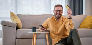 Porträt eines fröhlichen Mannes mit dem revolvierenden Kredit und eine Kreditkarte, der im heimischen Wohnzimmer sein Mobiltelefon benutzt.