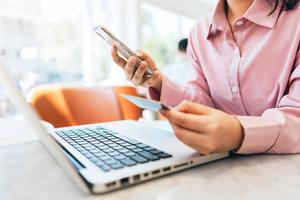 Eine Frau sitzt am Laptop und hält ein Smartphone und eine Bankkarte in der Hand