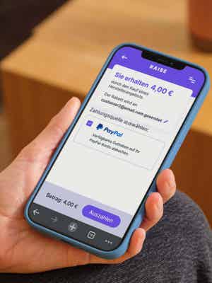 Person hält ihr Mobiltelefon mit geöffneter RAI$E-Anwendung; PayPal als Auszahlungsmethode ausgewählt