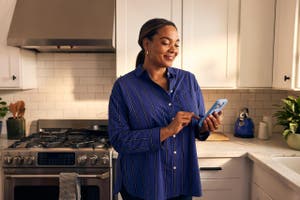 In a kitchen, a woman holds a cell phone, engaged in conversation while surrounded by cooking utensils and ingredients.