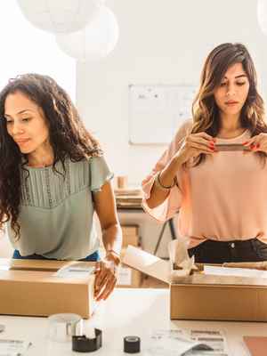 2 business women packing boxes with clothing orders; 1 woman is using her laptop to update their online store