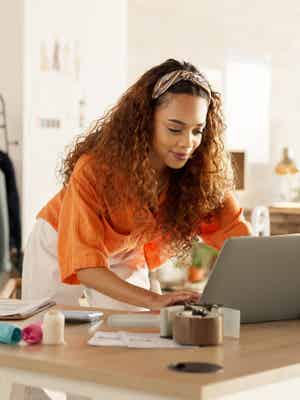 Smiling businesswoman in vibrant orange shirt and white pants applying for a PayPal Working Capital loan on her laptop