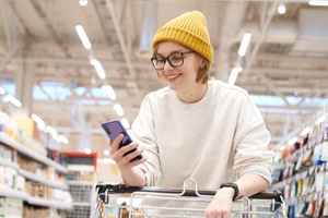 A smiling woman looking at her smartphone while holding onto a grocery cart.