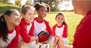 Four young girls in white and red baseball shirts kneeling on the grass