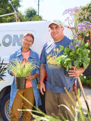 2 happy landscaping business owners; 1 is holding an ornamental grass, 1 is holding a large flowering plant