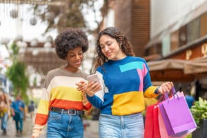 A woman holds shopping bags and shows her friend her phone.