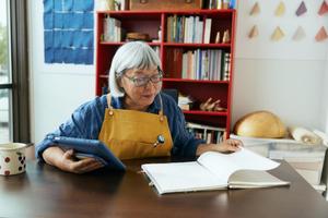 An older woman seated at a table, engaged with a tablet in one hand and a book in the other, showcasing her interests.